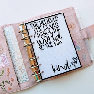 May include: A light pink leather-effect planner with a six-ring metal binder mechanism. The white insert features the black text: SHE BELIEVED SHE COULD CHANGE THE WORLD SO SHE WAS kind, accompanied by two small heart icons at the bottom right.