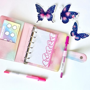 May include: A pastel-colored planner is open to a dotted page with a pink heart border. Three butterfly stickers and two pink pens are on a white surface. A card with stickers is in the planner pocket.
