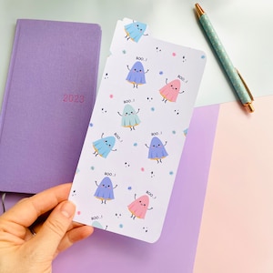 May include: A planner with a 2023 date and a decorative bookmark featuring cartoon ghosts saying "BOO!". The bookmark has a white background with colorful ghosts and stars. A pen with a gold tip rests on the background.