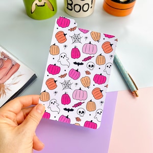 May include: A Halloween-themed planner insert with a white background. The design includes pink and orange pumpkins, ghosts, spiderwebs, bats, and skulls. A hand holds the insert, with a pen and Halloween mugs visible in the background.