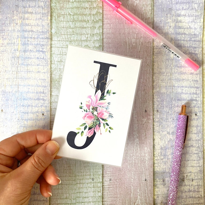 May include: A white card with a black letter J decorated with pink flowers and gold accents.