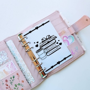 Può includere: Un'agenda in pelle rosa con una pagina bianca aperta. La pagina presenta un'illustrazione in bianco e nero di una pila di libri con una tazza di caffè. Ci sono piccoli cuori neri sparsi intorno all'illustrazione.