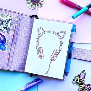 May include: A lilac planner featuring a cat ear headphone design, open to show a card and stickers. A blue pen and colourful stickers are also visible. The background is a mix of lilac and blue.
