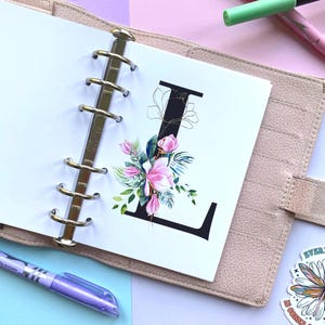 May include: A beige ring-bound planner featuring a white page with a large black letter L. The letter is decorated with a vibrant floral illustration of pink magnolia flowers, green leaves, and thin gold line art accents. A purple pen lies nearby.
