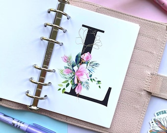 May include: A beige ring-bound planner featuring a white page with a large black letter L. The letter is decorated with a vibrant floral illustration of pink magnolia flowers, green leaves, and thin gold line art accents. A purple pen lies nearby.