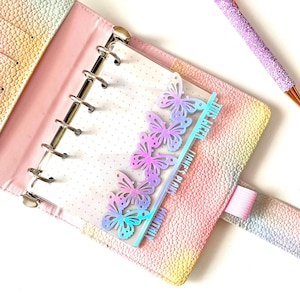 May include: A pastel-coloured planner with a butterfly-themed page marker. The marker is a clear, iridescent plastic with butterfly cutouts and text that reads "This Week", "Liane's Plans", and "Monthly". A glittery purple pen rests on the side.