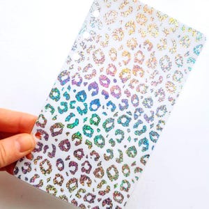 May include: A rectangular translucent plastic planner dashboard featuring a repeating holographic leopard print pattern. The surface displays a shimmering, multi-colored effect across the animal spots, and the left edge includes a vertical row of circular punched holes for binder insertion.