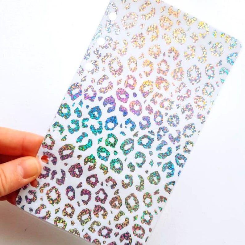 May include: A rectangular translucent plastic planner dashboard featuring a repeating holographic leopard print pattern. The surface displays a shimmering, multi-colored effect across the animal spots, and the left edge includes a vertical row of circular punched holes for binder insertion.