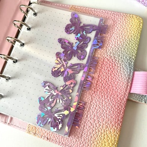 May include: A planner with a pink, textured cover and silver rings. A clear page with purple butterfly cutouts and the word "MONTHLY PLAN" is inserted. The planner is open, revealing a dotted page.