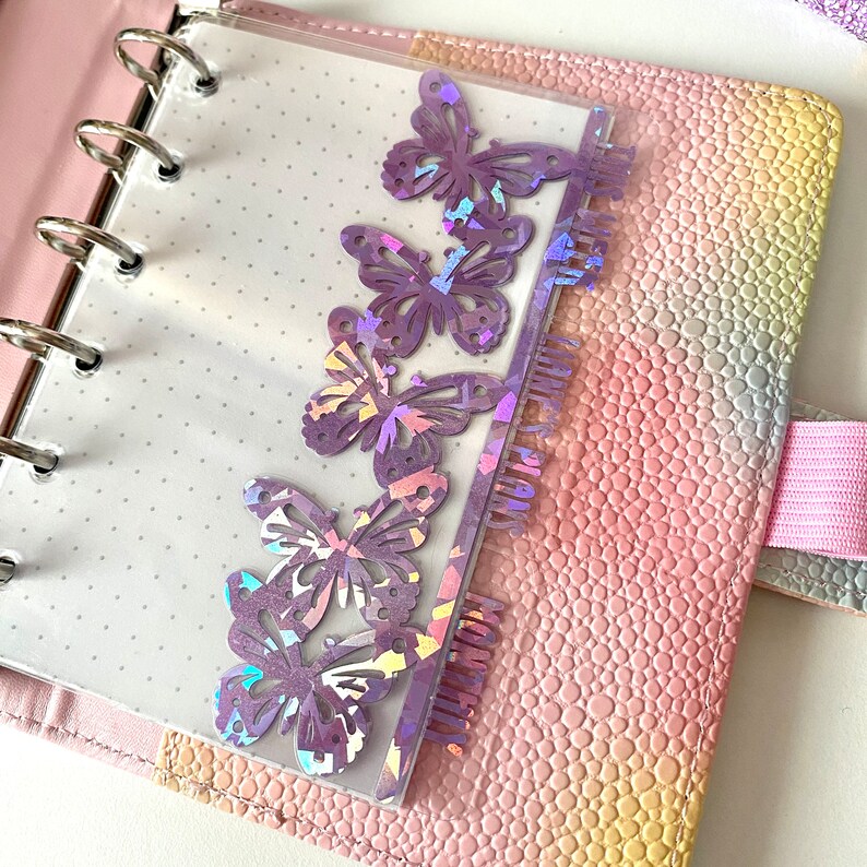 May include: A planner with a pink, textured cover and silver rings. A clear page with purple butterfly cutouts and the word "MONTHLY PLAN" is inserted. The planner is open, revealing a dotted page.