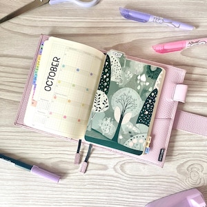 May include: A light pink leather planner cover holding an open planner with a monthly calendar page for October and a decorative green forest-themed page marker labeled TODAY. The planner features colorful star stickers and small index tabs.