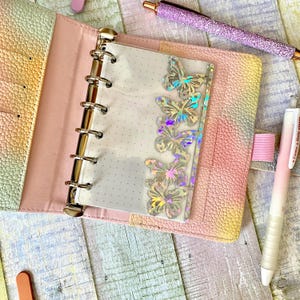 May include: Open pastel-colored planner with a clear butterfly-adorned divider. The planner is open, revealing a dotted page. A glitter pen and a pink and white pen are also visible, suggesting stationery or organizational supplies.