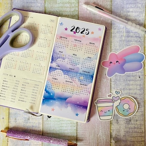 Può includere: Un calendario 2025 con uno sfondo ad acquerello di nuvole e stelle. Il calendario è aperto a luglio e agosto. Ci sono due adesivi sulla pagina, uno di una stella rosa e blu sorridente e l'altro di una tazza di caffè e un donut.