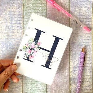 May include: A white card with a black letter H and pink floral design. The card has six holes on the left side for a binder.