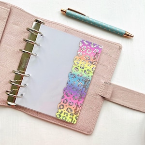 May include: A pink leather planner is open to a blank page, with a rainbow leopard print divider. A teal pen with a gold tip rests on the white surface. The divider has the words "Today", "Week", and "Month".