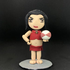 Puede incluir: Una figura artesanal de una mujer con cabello negro, vistiendo un top de bikini rojo y blanco y una falda roja. Ella sostiene una pelota blanca y roja en su mano derecha.