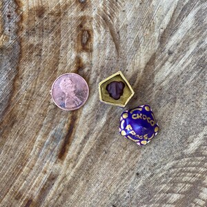 May include: A copper penny next to a small, gold-colored, geometric container with a dark brown interior. A wrapped, purple candy with gold lettering that reads "Choco Frogs" is also present. The items are on a wooden surface.