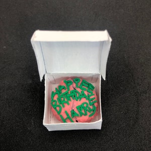 Può includere: Una torta in miniatura rotonda di colore rosa con scritta in glassa verde che recita "HAPPEE BIRTHDAE HARRY" all'interno di una piccola scatola di cartone bianca aperta. La torta ha una superficie ruvida e la scritta verde è disposta su tre righe.