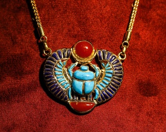 May include: Gold-toned necklace featuring an Egyptian scarab pendant. The pendant has a turquoise scarab beetle at the center, with blue and gold wings, and red accents. The necklace is displayed against a red background.