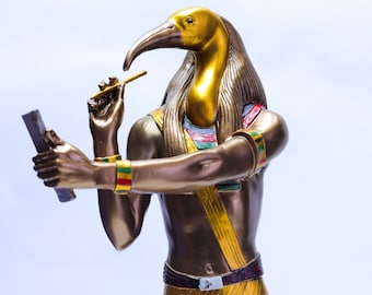 Könnte beinhalten: Eine goldene und bronzene Statue des ägyptischen Gottes Thoth, dargestellt als Mann mit dem Kopf eines Ibis. Er hält einen Stab in seiner rechten Hand und eine Schriftrolle in seiner linken Hand.