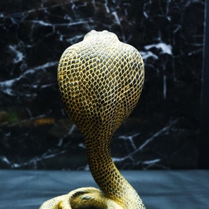 Op de afbeelding: Een gouden cobra-beeld met een gedetailleerd, getextureerd oppervlak. Het beeld is geposeerd met zijn kop omhoog en zijn lichaam opgerold.