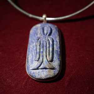 Op de afbeelding: Een zilveren ketting met een lapis lazuli hanger met een gegraveerd Egyptisch symbool. De hanger is ovaalvormig en heeft een donkerblauwe kleur met gouden vlekken.