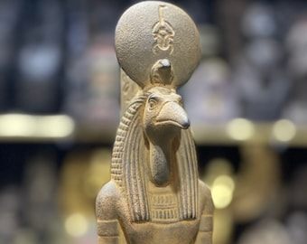 Könnte beinhalten: Detaillierte Statue in Bronzefarbe einer ägyptischen Gottheit. Die Figur hat einen vogelartigen Kopf, eine hohe Kopfbedeckung und ist mit aufwendigen Schnitzereien verziert. Die Statue steht aufrecht, mit Armen an den Seiten, vor einem verschwommenen Hintergrund.