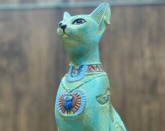May include: A turquoise blue cat statue with intricate details. The statue has a gold collar and a scarab beetle design on its chest. The cat is sitting upright with its head tilted up.