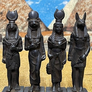 May include: Four black statues of Egyptian deities, each with a unique headpiece and standing on a square base. The statues are made of a dark, textured material and have a slightly weathered appearance.