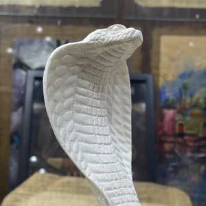 Op de afbeelding: Witte keramische sculptuur van een cobra met zijn kap omhoog. De sculptuur is gedetailleerd met schubben en een textuur.