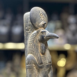 Puede incluir: Estatua detallada y desgastada del dios egipcio Thot, con cabeza de pájaro y tallas intrincadas. La estatua gris presenta un disco solar y jeroglíficos, mostrando el arte del antiguo Egipto.