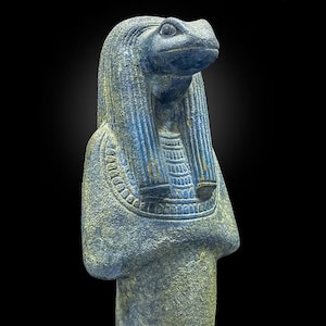 以下が含まれることがあります： カエルの頭を持つ人物の精巧な彫刻。古代エジプトの神である可能性が高い。像は主に青色で金色のアクセントがあり、頭飾りと襟に複雑なディテールが施されている。背景は黒色です。