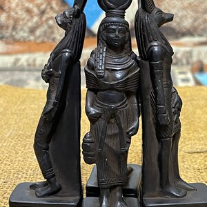 May include: Three black figurines of Egyptian deities. The center figure is a woman with a tall headdress, the figure on the left is a jackal-headed god, and the figure on the right is a dog-headed god.