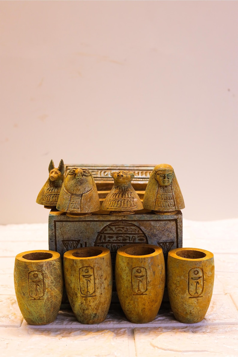 May include: A set of five small, green and brown ceramic cups with Egyptian hieroglyphics. The cups are arranged in a row on a white tiled surface. The cups are in front of a closed wooden box with a green and brown design and Egyptian hieroglyphics.