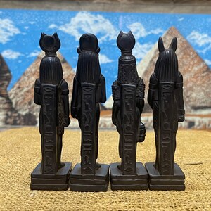 May include: Four black Egyptian god statues with hieroglyphics on the front. The statues are standing on a brown surface.