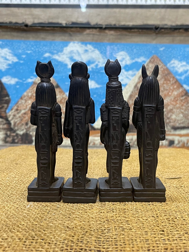 May include: Four black Egyptian god statues with hieroglyphics on the front. The statues are standing on a brown surface.