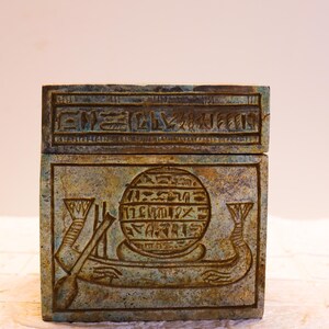 May include: A small, square, green and brown box with hieroglyphics carved into the top and front. The front features a circular design with hieroglyphics and a winged scarab beetle.
