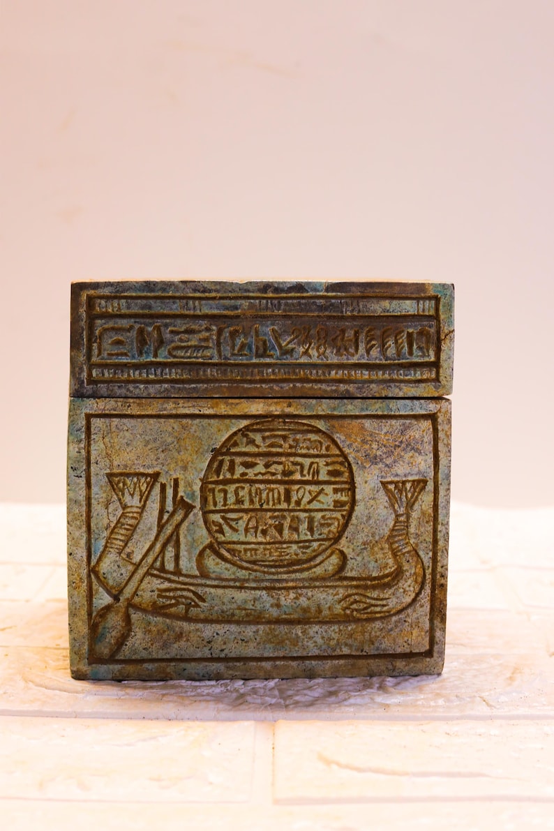 May include: A small, square, green and brown box with hieroglyphics carved into the top and front. The front features a circular design with hieroglyphics and a winged scarab beetle.