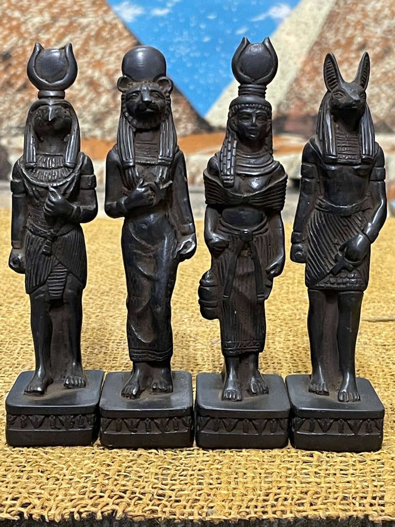 May include: Four black statues of Egyptian deities, each with a unique headpiece and standing on a square base. The statues are made of a dark, textured material and have a slightly weathered appearance.