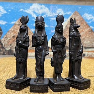 May include: Four black figurines of Egyptian gods and goddesses. The figures are standing on small black bases. The figures are all wearing traditional Egyptian clothing and jewelry.