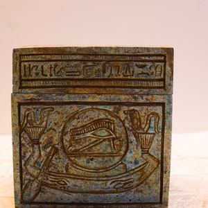 May include: A small, rectangular box made of green stone with intricate carvings. The box has a lid with hieroglyphics on it. The carvings on the box depict an eye with a winged sun disk above it.