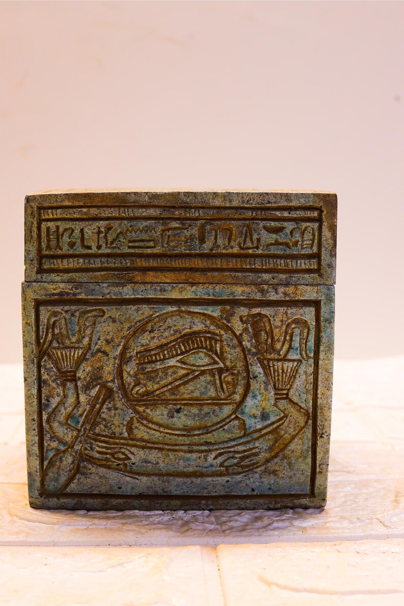 May include: A small, rectangular box made of green stone with intricate carvings. The box has a lid with hieroglyphics on it. The carvings on the box depict an eye with a winged sun disk above it.