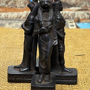 May include: Black resin statue of three Egyptian deities. The central figure is a cat-headed god holding a sphere. The figures stand on separate bases with decorative carvings.