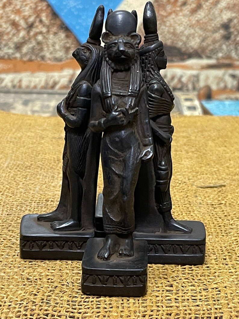 May include: Black resin statue of three Egyptian deities. The central figure is a cat-headed god holding a sphere. The figures stand on separate bases with decorative carvings.
