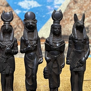 May include: Four black figurines of Egyptian deities, each with a distinct headpiece and detailed clothing. The figurines are standing on a brown surface.