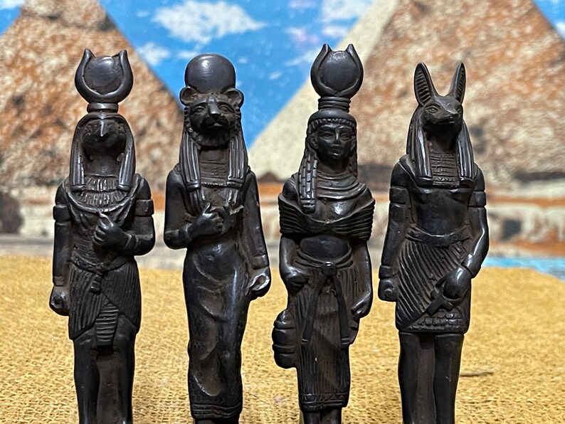 May include: Four black figurines of Egyptian deities, each with a distinct headpiece and detailed clothing. The figurines are standing on a brown surface.