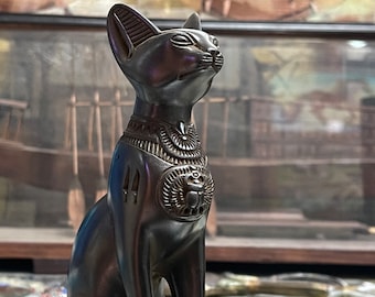 May include: A dark bronze cat statue in a seated pose, with detailed carvings. The statue features an Egyptian-style collar and chest designs, including a scarab beetle. The cat's ears are perked, and it looks upwards.