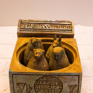 May include: A small, square, wooden box with a hinged lid. The box is decorated with hieroglyphics and contains four small, carved figures of Egyptian deities. The figures are made of a light brown material and have intricate details.