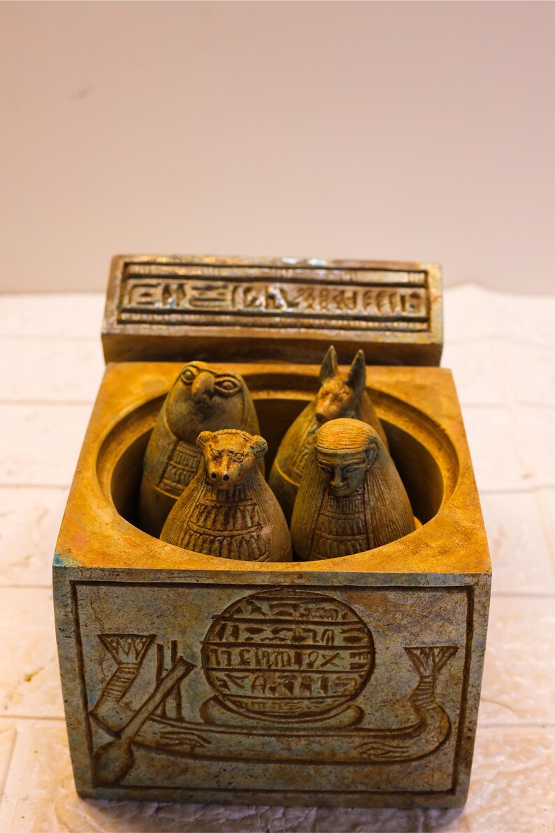 May include: A small, square, wooden box with a hinged lid. The box is decorated with hieroglyphics and contains four small, carved figures of Egyptian deities. The figures are made of a light brown material and have intricate details.