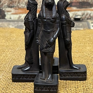May include: Three black figurines of Egyptian gods, each standing on a square base with intricate carvings. The god in the center is Anubis, the jackal-headed god of the dead, with a headdress and a staff. The god on the left is a woman with a tall headdress and a staff. The god on the right is a man with a falcon head and a staff.